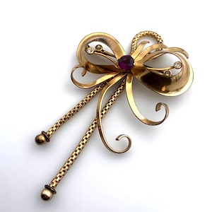 May include: A vintage gold-toned brooch in a bow design, featuring a central purple gemstone and smaller clear stones. Two chain-like elements extend downward, each ending in a small, round detail. The brooch is set against a white background.