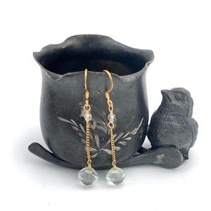 May include: A pair of gold-colored earrings with clear, faceted crystal beads. The earrings hang from a gold chain and are displayed next to a dark gray, decorative pot and a small bird figurine.