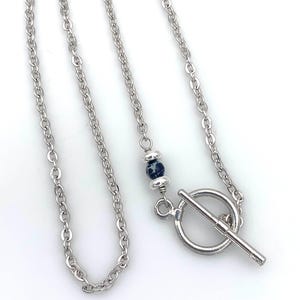 May include: Silver-toned necklace with a toggle clasp. The chain features a small, dark blue and black speckled bead. The necklace is displayed on a white background.