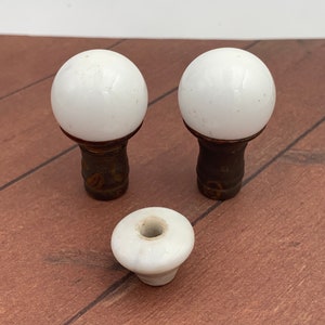 May include: Three antique white porcelain cabinet knobs with brass bases. Two knobs are spherical, and one is a flattened disc shape. The brass bases have an aged, dark finish. The knobs are on a brown wooden surface.