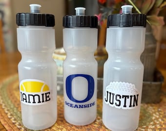 May include: Three translucent plastic water bottles with black screw-on lids and pull-up spouts. The left bottle features a yellow tennis ball graphic and the name JAMIE. The center bottle displays a blue letter O and the text OCEANSIDE. The right bottle shows a white football graphic and the name JUSTIN.
