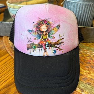May include: A black trucker hat with a pink top panel featuring a watercolor illustration of a fairy. The fairy has colorful wings, hair, and clothing, and is sitting on a branch. The hat has a mesh back.