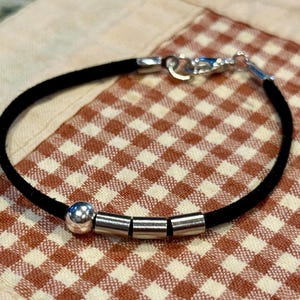 May include: A black suede bracelet with silver-toned metal detailing. The bracelet has a clasp fastening and includes three cylindrical beads and one spherical bead. It is shown on a red and white checked pattern.
