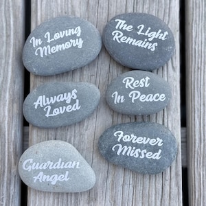 May include: Six smooth, grey river stones featuring white engraved text. The stones display the phrases In Loving Memory, The Light Remains, Always Loved, Rest In Peace, Forever Missed, and Guardian Angel in a cursive, white script font.