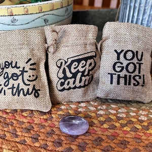 May include: Three small burlap drawstring bags with black text that reads "You got this!", "Keep calm", and "You got this!". A small, oval, purple stone sits in front of the bags. The bags are on a woven, multi-colored surface.