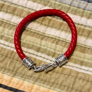 May include: A red leather bracelet with silver-toned clasps and decorative accents. The bracelet is braided and has a simple design.
