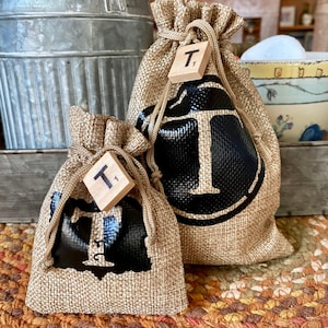May include: Two small burlap bags with black painted letter "T" on the front. The bags are tied with twine and have a small wooden tile with the letter "T" on them.