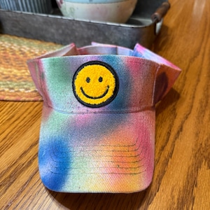 May include: A tie-dye visor in shades of blue, pink, yellow, and green. The visor features a yellow smiley face patch. The visor is designed to shield the eyes from the sun.