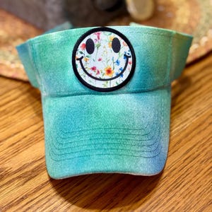 May include: A tie-dye visor in shades of blue and green. The visor features a circular patch with a smiley face design filled with colourful flowers. The visor is made of a soft, textured fabric.