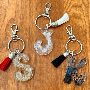 May include: Three keychains with silver hardware and letter-shaped pendants. The letters are filled with glitter and paired with colorful tassels. The letters are "S", "J", and "K". The tassels are red, white, and black.