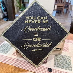 May include: A black glittery graduation cap topper featuring gold lettering that reads: YOU CAN NEVER BE Overdressed OR Overeducated OSCAR WILDE. The square topper is bordered by a gold rhinestone trim and has a circular hole in the centre.