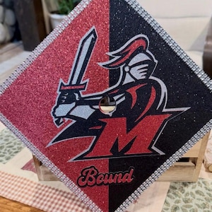 May include: A square graduation cap topper featuring a split red and black glitter background. It displays a silver and black knight mascot holding a sword, a large red letter M, and the word Bound in script. The edges are lined with rhinestones.