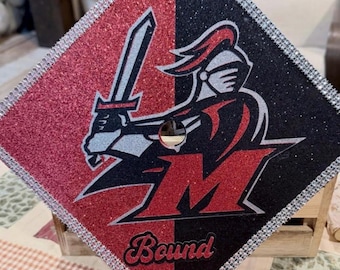 May include: A square graduation cap topper featuring a split red and black glitter background. It displays a silver and black knight mascot holding a sword above a large red letter M and the word Bound in script. The edges are lined with rhinestones.