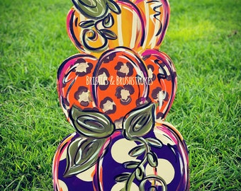 Pode incluir: Uma pilha de três abóboras de madeira pintadas. A abóbora de cima é laranja e branca listrada, a abóbora do meio é laranja e preta com estampa de leopardo, e a abóbora de baixo é roxa com bolinhas brancas. As abóboras têm folhas e caules verdes.