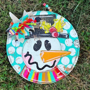 May include: A round, turquoise and white polka dot door hanger with a painted snowman face. The snowman has a black hat with a green holly sprig, a black and orange carrot nose, and black eyes. The bottom of the hanger has a colourful striped design.