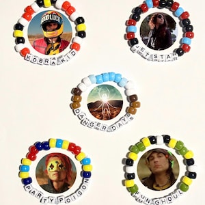 Puede incluir: Cinco pulseras de cuentas coloridas con imágenes circulares de personas. Cada pulsera tiene una combinación de colores diferente y un nombre escrito con cuentas de letras blancas. Los nombres incluyen "KOBRA KID", "JET STAR", "DANGER DAY", "PARTY POISON" y "FUN GHOUL".