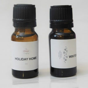 May include: Two amber glass essential oil bottles with black caps. One bottle is labeled "HOLIDAY HOME" and the other is labeled "WINTER". The bottles have white labels with black text and a small logo.