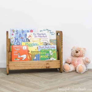 Könnte beinhalten: Ein Bücherregal aus Holz mit einer Auswahl an Kinderbüchern, darunter "Super Happy Magic Fun", "To Market, To Market", "Piggies", "Duck Duck Goose" und "Goodnight Moon". Ein rosa Teddybär sitzt auf dem Boden vor dem Bücherregal.