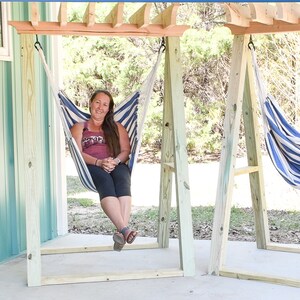 May include: Two wooden hammock stands with blue and white striped hammocks hanging from them. A person is sitting in one of the hammocks.