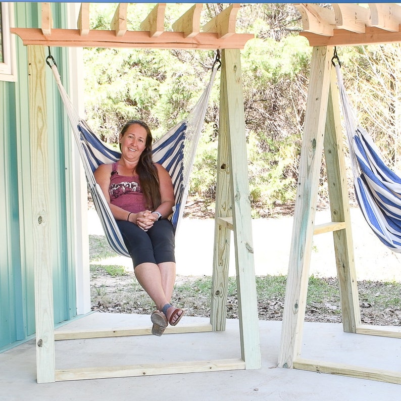 May include: Two wooden hammock stands with blue and white striped hammocks hanging from them. A person is sitting in one of the hammocks.