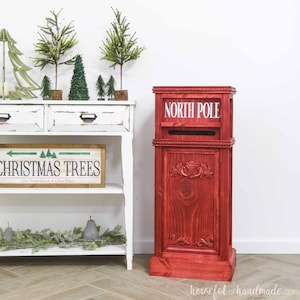 May include: A red wooden North Pole mailbox with decorative carvings. The mailbox has a slot for letters and the words "NORTH POLE" in white. The mailbox is part of a Christmas-themed display.