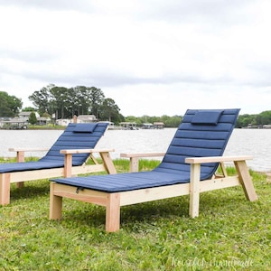 May include: Two wooden lounge chairs with blue cushions and headrests. The chairs are on a grassy lawn with a lake in the background.
