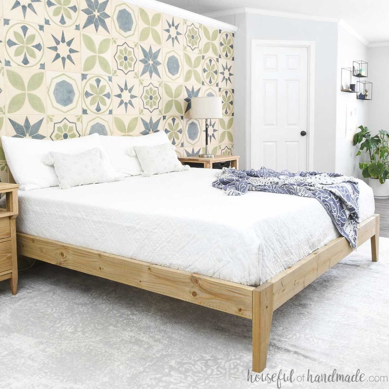 May include: A wooden bed frame with a white mattress and a blue blanket. The headboard is against a wall with a patterned wallpaper featuring blue and green geometric shapes.