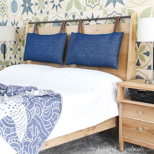 May include: A wooden bed frame with a headboard featuring two blue pillows suspended by leather straps. The bed is made up with a white comforter and a blue patterned throw blanket. A wooden nightstand with a lamp is on the right.