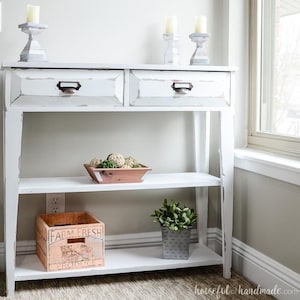May include: White distressed console table with two drawers and a lower shelf. The table is decorated with candles, decorative spheres, a wooden crate with "FARM FRESH" text, and a metal pot with greenery.