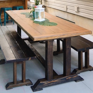 May include: A wooden table with a natural wood finish and two matching benches. The table has a farmhouse style with a thick, rectangular top and a sturdy base. The benches are made of dark stained wood and have a simple design.