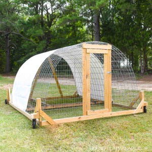 Puede incluir: Un gallinero móvil de madera con estructura de malla metálica y cubierta blanca. El gallinero tiene una puerta y está montado sobre ruedas. Se encuentra sobre césped, con árboles al fondo.