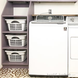 May include: A white washing machine and dryer are in a laundry room with a purple shelf unit. The shelf unit has three white baskets on it. The floor is tiled with brown and white tiles.