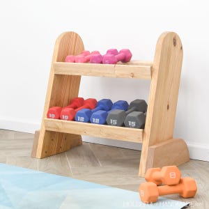 Op de afbeelding: Een houten halterrek met diverse gekleurde dumbbells. Roze, rode, blauwe en grijze dumbbells zijn netjes op het rek geplaatst. Twee oranje dumbbells van 3,6 kg liggen op de vloer. Het rek is gemaakt van lichtgekleurd hout.