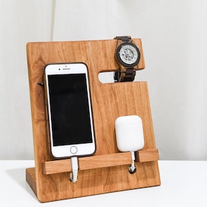 May include: A wooden phone and watch stand with a slot for a phone, a watch, and a small device like AirPods. The stand is made of light brown wood and has a natural finish.