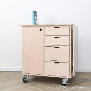 May include: A light-colored wooden cabinet with four drawers and a door, mounted on casters. Two books rest on top. The cabinet is made of plywood and has a simple, modern design. The cabinet is approximately 30 inches tall.