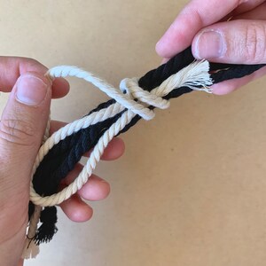 May include: Two ropes, one black and one white, are tied together in a knot. The ropes are held in place by two hands.