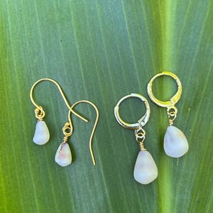 以下が含まれることがあります： 涙型のオフホワイトのペンダントが付いた2組のイヤリング。片方はゴールドカラーのフックピアス、もう片方はシルバーカラーのフープクロージャーです。イヤリングは緑の葉の上に展示されています。