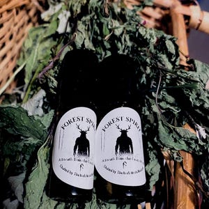 Puede incluir: Dos botellas oscuras con etiquetas blancas. Las etiquetas dicen "FOREST SPIRIT" y "A Breath from the Forest". Las etiquetas presentan una silueta de una figura con cuernos. Las botellas están rodeadas de hojas verdes.