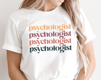 May include: White t-shirt with the text "psychologist" repeated five times in different colors. The text is stacked vertically, with each word in a different color. The colors are orange, pink, purple, gray, and dark gray. The t-shirt is being worn by a person sitting on a chair. The background is a white wall. The image also includes a blue circle with the text "little cutes studio" and "MORE COLORS & STYLES" in white text.