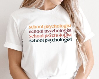 May include: White t-shirt with the text "school psychologist" repeated four times in different colors. The text is stacked vertically. The shirt is being worn by a person sitting on a chair. A blue circle with the text "little cutes studio" and "Colors & Styles" is in the lower right corner of the image.