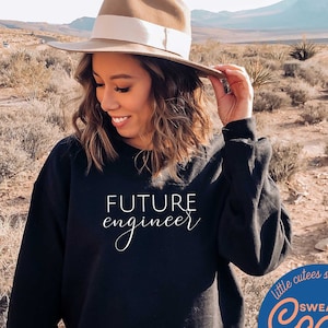 May include: A black sweatshirt with the text "FUTURE engineer" in white lettering. The sweatshirt is being worn by a person in a desert setting. The image also includes a blue circle with the text "little cutes studio Cozy SWEATSHIRT HOODIES" in white lettering.
