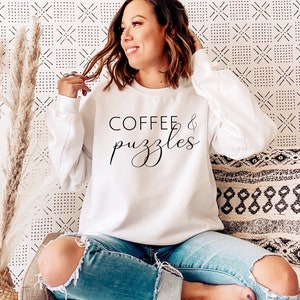 May include: A white sweatshirt with the text "COFFEE & puzzles" in black lettering. The sweatshirt is being worn by a person sitting on a couch.