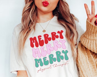 May include: White t-shirt with a festive design featuring the word "Merry" repeated three times in red, pink, and green. The text "physical therapist" is written below the word "Merry".