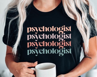 May include: A black t-shirt with the word "psychologist" repeated four times in different colors. The text is stacked vertically and the colors are pink, peach, light blue, and teal. The person wearing the shirt is holding a white mug.