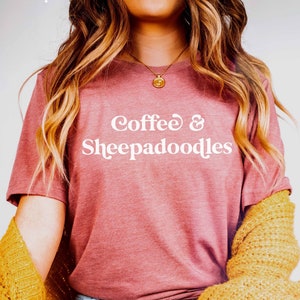May include: A woman wearing a mauve t-shirt with the text "Coffee & Sheepadoodles" in white lettering. She is wearing blue jeans and a yellow cardigan.