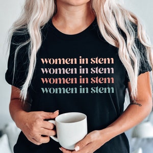 May include: A black t-shirt with the text "women in stem" repeated four times in different colors. The text is in a bold, sans-serif font. The shirt is being worn by a person with blonde hair.