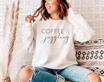 May include: A white sweatshirt with the text "Coffee & Juggling" in black lettering. The sweatshirt is being worn by a person sitting on a couch.