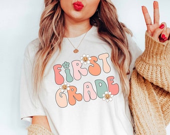 May include: A white t-shirt with the text "FIRST GRADE" in colorful, retro-style lettering with daisies. The shirt is being worn by a person with long blonde hair and a tan cardigan.