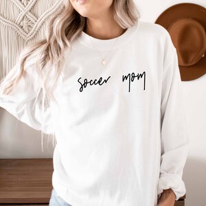 May include: White long-sleeved sweatshirt with the text "Soccer Mom" written in black cursive script.