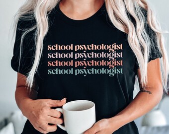 May include: A black t-shirt with the text "school psychologist" repeated four times in different colors. The text is stacked vertically, with each line in a different color: pink, orange, blue, and green. The person wearing the shirt is holding a white mug.